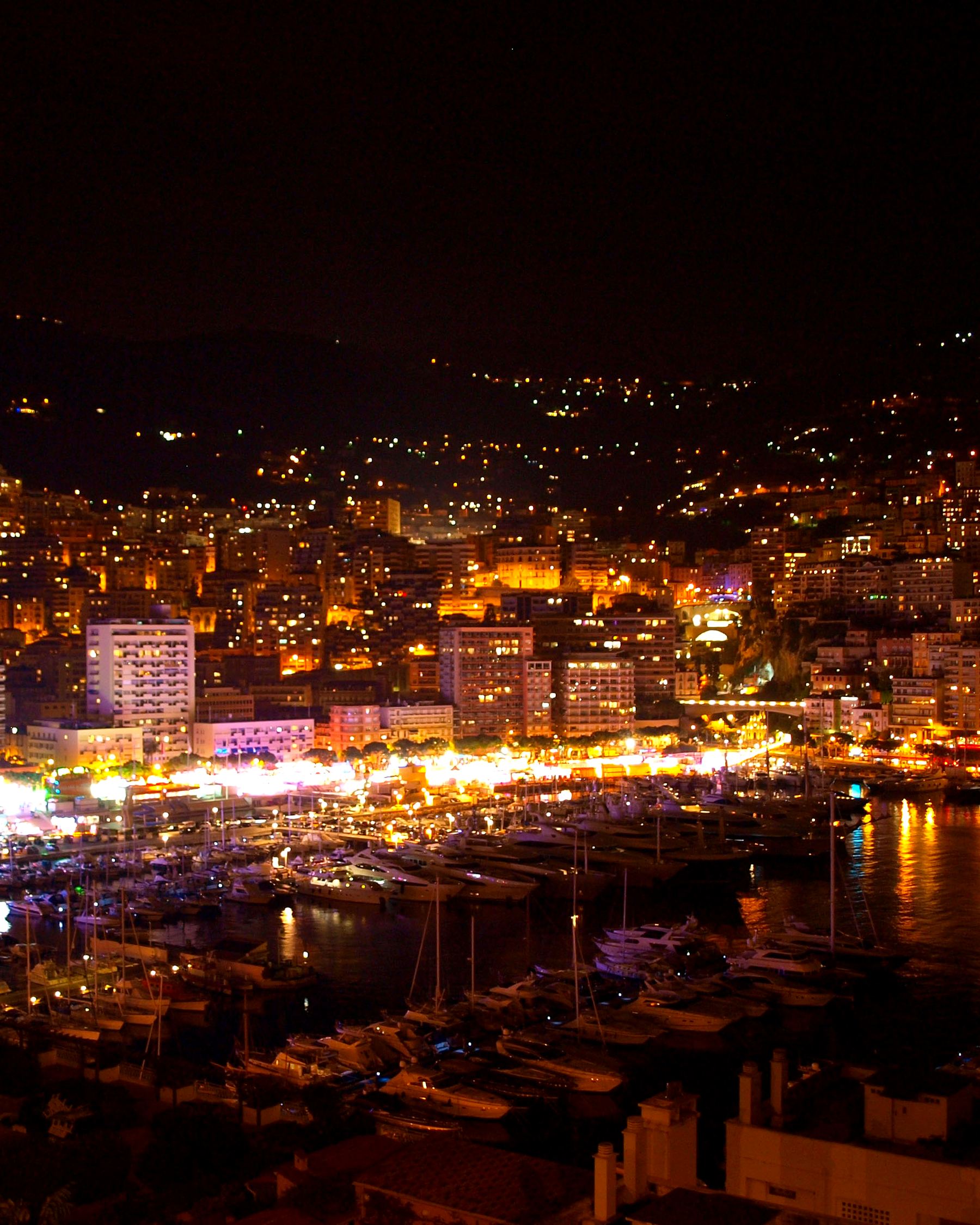 Monaco at night from above: the harbour, the lit yachts and the city climbing the hill behind.