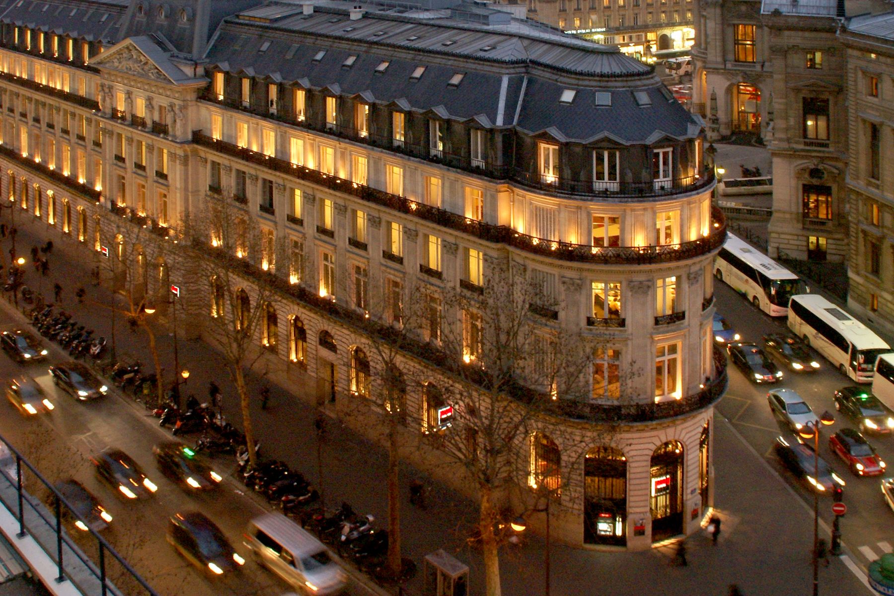 A Paris corner building at dusk, its windows lit, traffic passing below.