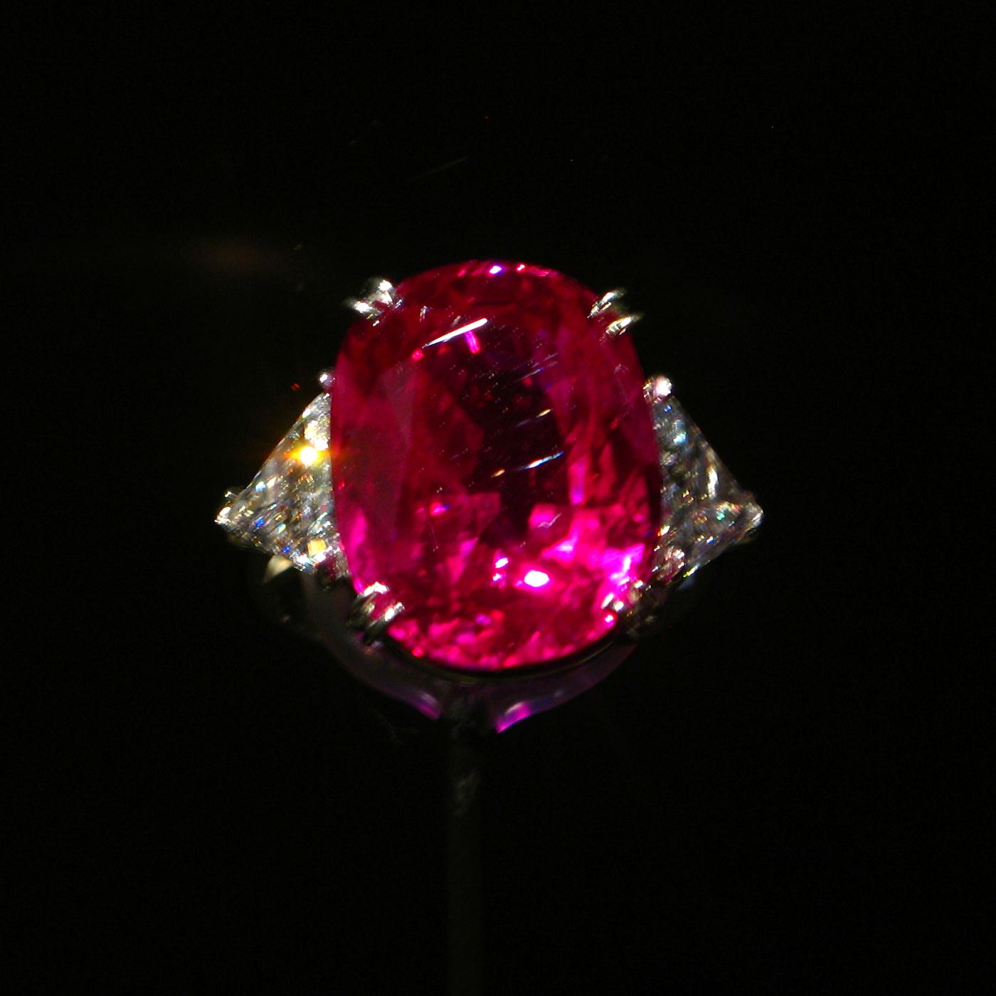 An oval pink-red ruby set between two diamonds, glowing against black.